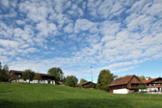 grüne Wiese mit Holzhäusern und blauer Himmel mit weißen Wölkchen