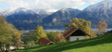 grüne Wiese im Hintergrund Berge und dazwischen ein See und Bäume und ein Haus